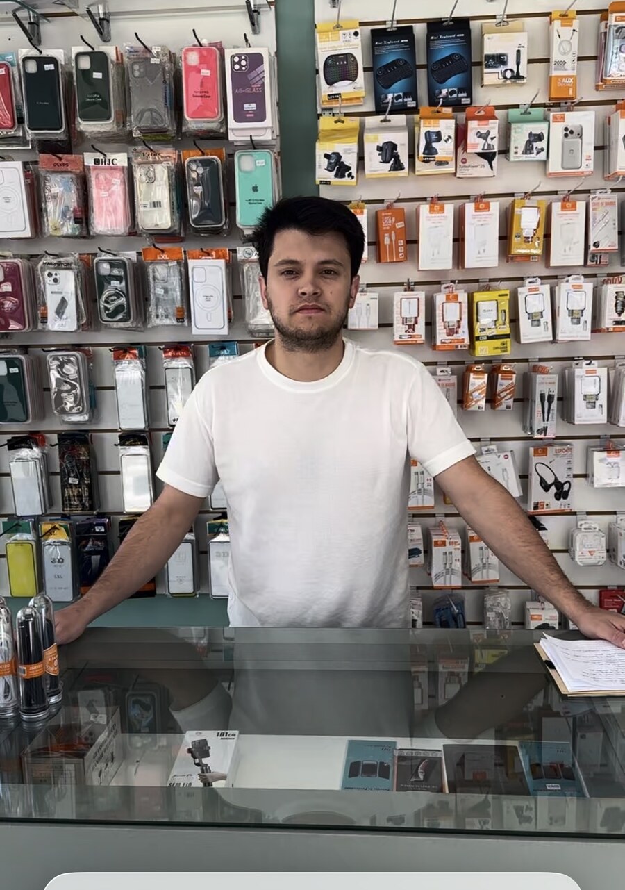 Daniel Pinheiro atrás do balcão da Conserta CN, com os braços apoiados e acessórios da loja ao fundo
